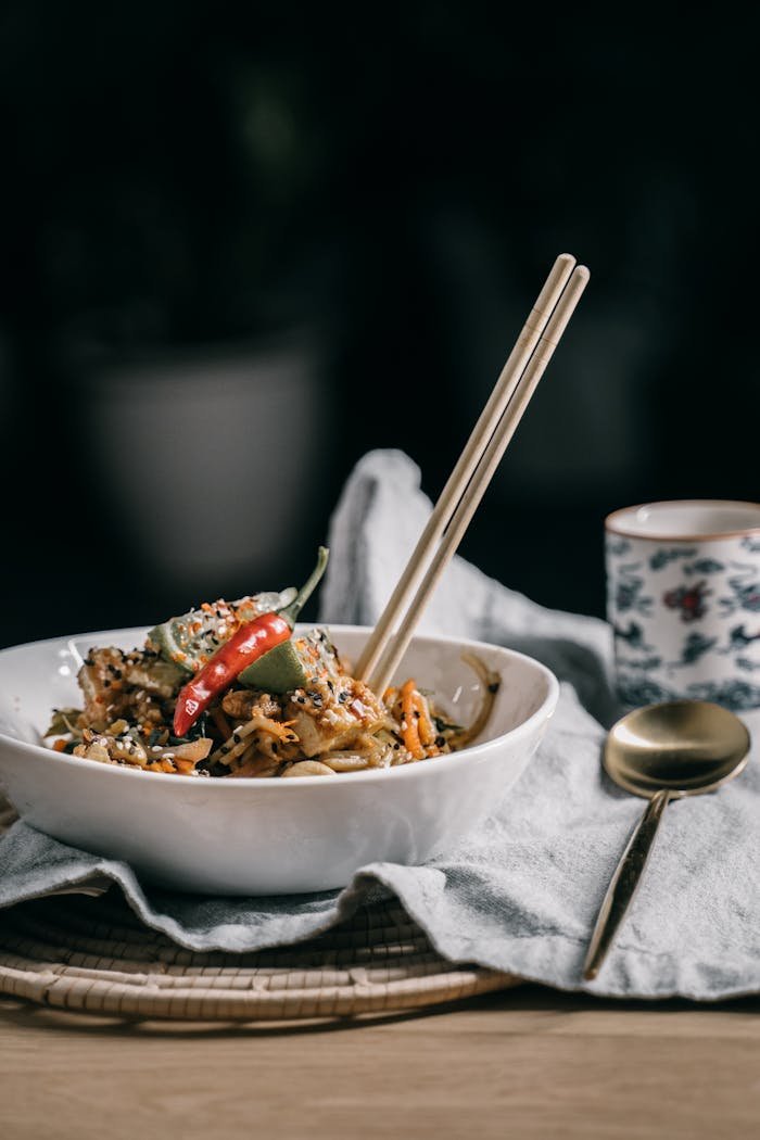A vibrant bowl of Asian noodles with chopsticks, adorned with colorful vegetables and spices, served alongside a cup of tea.